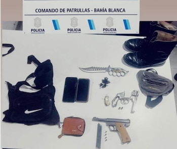 Armas de fuego, un cuchillo, teléfonos celulares, una billetera, bolsos y un par de botas sobre una mesa ante un cartel de la policía