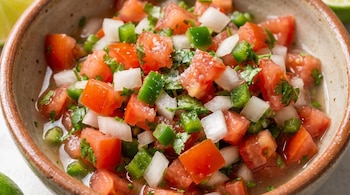 Primer plano de salsa pico de gallo en un cuenco rústico, con trozos visibles de tomate rojo, cebolla blanca, jalapeño verde y cilantro picado, con cristales de sal.