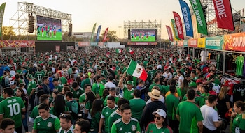 Multitud de personas con camisetas verdes de fútbol. Varias pantallas gigantes muestran un partido. Banderas de México y puestos de venta de ropa.