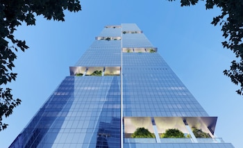 Rascacielos de cristal alto con diseño escalonado y terrazas verdes. Cielo azul claro. Hojas de árbol oscuro en las esquinas superiores