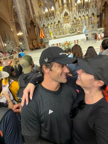 Un hombre y una mujer con gorras negras se abrazan y sonríen en el interior de una iglesia frente a un altar