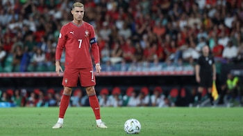 Cristiano Ronaldo vestido con el uniforme rojo número siete parado frente a un balón de fútbol sobre el césped de un estadio