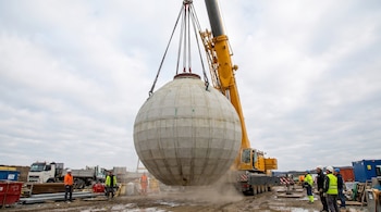 Una grúa amarilla levanta una esfera de hormigón masiva en un sitio de construcción. Seis trabajadores con equipo de seguridad observan la operación en terreno fangoso.