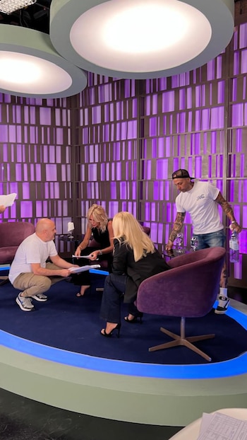 Cuatro personas, dos mujeres rubias y dos hombres, interactúan en un set de televisión con sillas moradas, fondo púrpura cuadriculado y luces de estudio