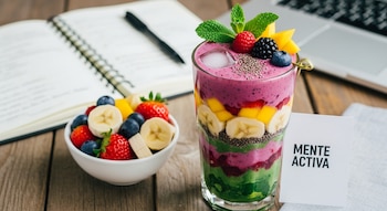 Vaso de batido de frutas en capas con hielo, menta, bayas, mango y chía, junto a un bol de frutas frescas, cuaderno y tarjeta "MENTE ACTIVA" en mesa de madera.