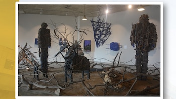 Interior de una galería de arte con dos figuras humanoides y una escultura abstracta, todas hechas de ramas secas y madera. Hay objetos azules y ramas en el suelo