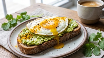 Rebanada de pan integral con aguacate, huevo frito con yema escurriendo, cúrcuma y pimienta en un plato, con cilantro y una taza de café.