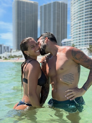 Un hombre con tatuajes besa en la mejilla a una mujer sonriente dentro del agua, con edificios altos en el fondo