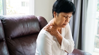 Una mujer de cabello oscuro corto y camiseta blanca está sentada en un sofá de cuero marrón. Sujeta su hombro izquierdo con la mano derecha, cerca de una ventana.