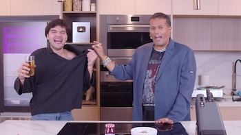 Dos hombres en una cocina. Joven con vaso sonríe. Hombre mayor sujeta su camisa. Refrigerador con texto, horno y premio YouTube plateado en el fondo