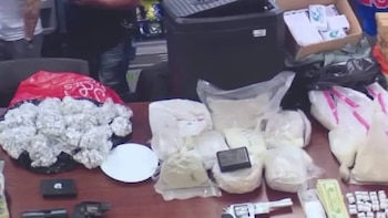 Una mesa cubierta con una gran cantidad de drogas, incluyendo paquetes individuales, varias armas de fuego, fajos de billetes y una báscula digital