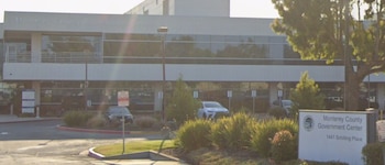 Edificio blanco con ventanas, un cartel con el texto Monterey County Government Center, coches estacionados, arbustos y árboles frondosos