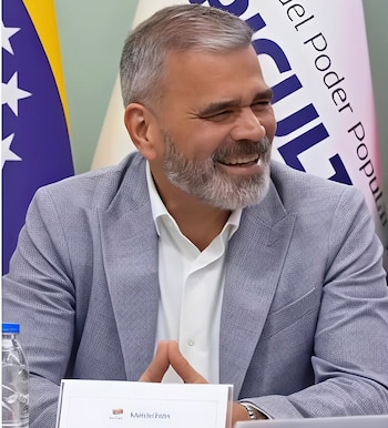 Hombre de barba y saco gris sonriente en una mesa con las manos juntas frente a dos banderas