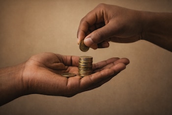 Primer plano de dos manos morenas de adultos. Una mano sostiene una moneda dorada a punto de colocarla sobre una pila de otras monedas en la palma de la otra mano abierta.