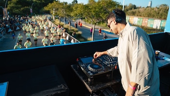 Vista aérea de un DJ con auriculares y gafas de sol, manejando una mesa de mezclas. Debajo, una multitud de corredores avanza por una calle arbolada