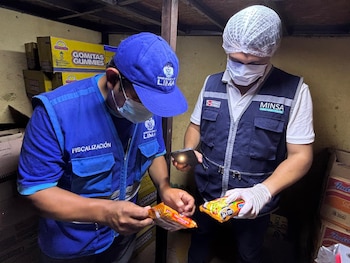 Dos personas con chalecos de fiscalización y mascarillas examinan empaques de comida frente a cajas de golosinas en un depósito
