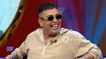 Dio's Sorrow riendo, con gafas de sol ovaladas doradas y una sudadera con capucha beige. Tiene tatuajes en cuello y brazos, y varios piercings. Fondo rojo y azul