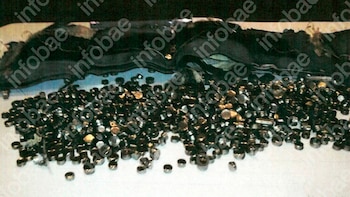 Primer plano de numerosos pellets pequeños, de color oscuro y metálico, esparcidos sobre una superficie clara con un material textil oscuro al fondo