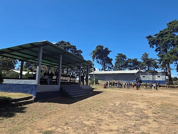 Escena al aire libre con escenario cubierto, personas sentadas y de pie, y un grupo en un campo. Edificios y árboles al fondo bajo cielo azul