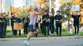 Corredor con atuendo morado, de espaldas y brazos levantados, pasa junto a una banda de música con instrumentos y una mujer con un cartel
