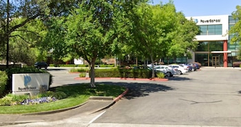 Edificio moderno de PowerSchool con fachada de vidrio y blanco. En el primer plano, un letrero de la empresa rodeado de plantas y un estacionamiento con varios autos