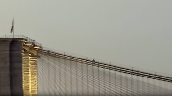 Parte superior de un puente de suspensión con cables, torre de piedra, una bandera y una silueta de persona en la pasarela