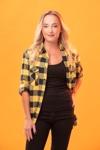 Una mujer rubia con cabello largo, vistiendo una camisa a cuadros amarilla y negra abierta sobre un top negro, posa sonriendo ligeramente contra un fondo naranja liso