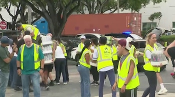 Grupo de personas con chalecos de seguridad amarillos cargan cajas de agua en un estacionamiento. Hay automóviles y un contenedor de carga en el fondo
