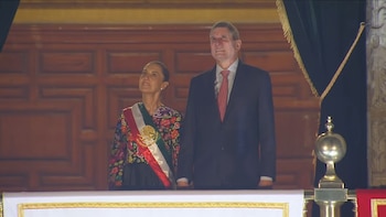 Una mujer con vestido bordado y banda tricolor junto a un hombre con traje oscuro observan desde un balcón