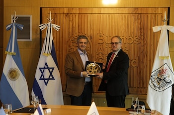 Dos hombres de pie intercambian una placa dorada en una sala con banderas de Argentina e Israel y una pared de madera con texto. Hay una mesa con vasos