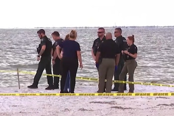 Un grupo de agentes policiales y personal de emergencia se reúne en una playa con el océano de fondo, donde una cinta amarilla delimita un área en la arena