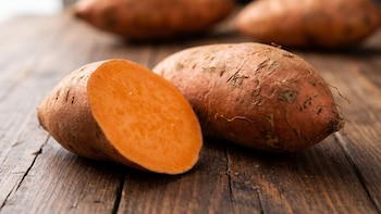 Una batata cortada por la mitad con el interior anaranjado y varias batatas enteras sobre una mesa de madera