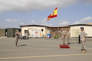 Militares con uniforme de camuflaje saludan frente a la bandera de España izada en un mástil en un recinto militar