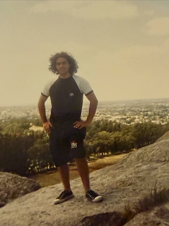 Un hombre joven de cabello rizado oscuro posa sobre una roca con las manos en las caderas, vista de un paisaje urbano arbolado al fondo