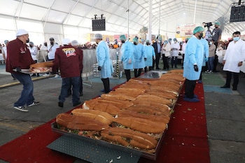 Varias personas vestidas con ropa protectora y otras en chaquetas rojas preparan una torta gigante en un evento bajo una carpa blanca