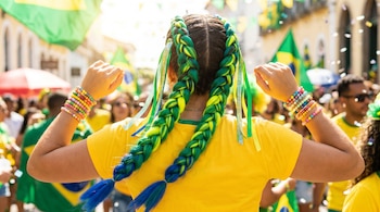 Imagen de la espalda de una persona con trenzas boxer verde esmeralda, amarillo neón y azul, camiseta amarilla y pulseras, en un evento con banderas de Brasil.