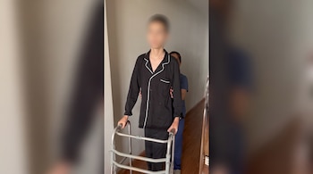Un joven en pijama negro camina con un andador metálico mientras una profesional de la salud lo sostiene por la espalda en un pasillo