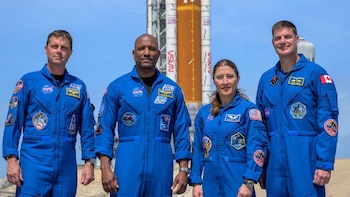 Cuatro astronautas en uniformes de vuelo azules posan frente a un cohete espacial blanco y naranja de la NASA. Cielo azul claro de fondo