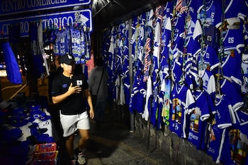 Un hombre camina junto a un puesto callejero con camisetas azules y blancas colgadas para la venta
