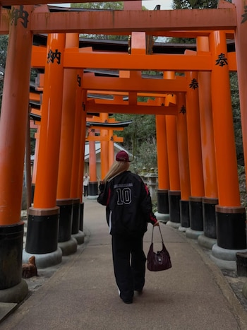 Vista trasera de Wanda Nara, con cabello rubio, gorra roja y chaqueta negra Balenciaga, caminando por un sendero entre numerosos torii naranjas