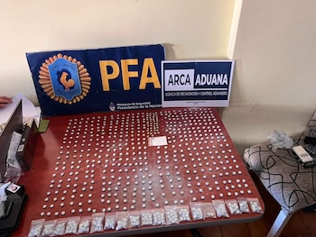 Una mesa de madera con cientos de pastillas blancas, algunas en bolsas, y dos carteles de Policía Federal Argentina y Agencia de Recaudación y Control Aduanero