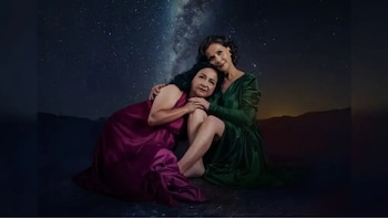 Dos mujeres sentadas con vestidos, una magenta y otra verde, bajo un cielo estrellado con la Vía Láctea y montañas difusas al fondo
