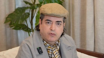El periodista Jaime Bayly viste una gorra beige, saco claro y bufanda a cuadros frente a una planta y cortinas