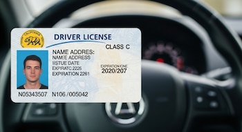 Primer plano de una licencia de conducir de California con la foto de un hombre, el sello del estado y el texto "DRIVER LICENSE", sobre un volante borroso de coche.