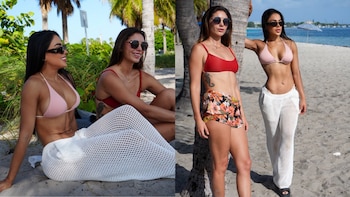 Dos mujeres con bikinis rosa y rojo posan en una playa arenosa con palmeras y el mar. Usan gafas de sol, pantalones de malla y shorts