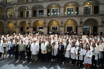 Gran grupo de profesionales de la salud, muchos con batas blancas, sosteniendo certificados en alto dentro de un amplio patio interior con múltiples arcos y balcones