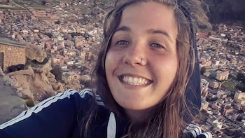 Primer plano de una joven sonriente con cabello castaño y sudadera oscura, un piercing en la nariz, y una extensa ciudad en la ladera de una montaña de fondo