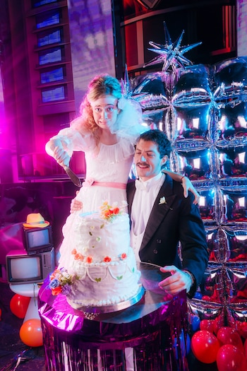 Una mujer con vestido blanco y velo sostiene un cuchillo junto a un hombre en esmoquin, frente a un pastel de bodas de varios pisos y globos