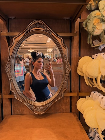 Mujer con orejas de Minnie Mouse negras y un top azul oscuro se toma una selfie en un espejo ovalado con marco dorado; al fondo, una tienda con orejas de Disney