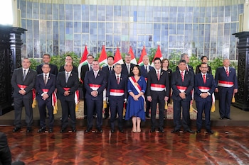 Grupo de diecisiete personas en traje formal posa en un salón. Dieciséis hombres con faja bicolor y una mujer en vestido azul con faja presidencial. Banderas de Perú al fondo